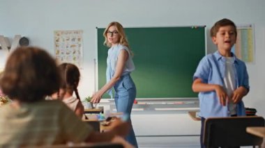 Kadın öğretmen modern sınıfa girerek derse başladı. Çocuk ilkokuluyla pozitif bayan eğitimci selamlaşması. Gözlüklü sarışın bayan öğretmen sınıftaki ilkokul öğrencileriyle konuşuyor..