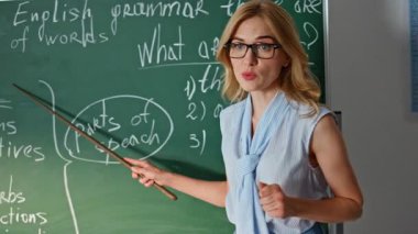 İngilizce dil bilgisi kurallarını açıklayan öğretmen sınıf tahtasında yakın plan metni işaret ediyor. Okul öğrencilerine ders veren profesyonel bir kadın. Gözlüklü gülümseyen bayan eğitmen çocuklarla konuşuyor..