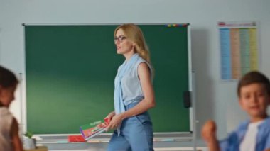 Öğretmen sınıfa giriyor ve grup ilkokul öğrencileriyle selamlaşıyor. Dost canlısı kadın sınıftaki meraklı çocuklarla konuşmaya başladı. Çocuklarla tanışan, gözlüklü, gülümseyen bayan eğitmen..