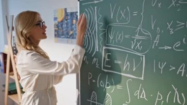 Öğretmen tahtaya fizik formülleri yazıyor. Bilinmeyen öğrencilere bilgi veriyor. Kadın eğitimci, tahtada matematik denklemleri öğretiyor. Okulda çalışan gözlüklü profesyonel bir öğretmen..