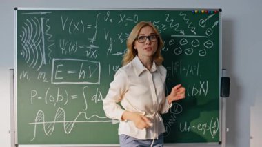 Bilinmeyen üniversite öğrencilerine matematik formüllerini açıklayan bir kadın. Gözlüklü gülümseyen öğretmen tahtaya matematiksel denklemler yazıyor. Okulda öğretmenlik yapan pozitif kadın eğitmen.
