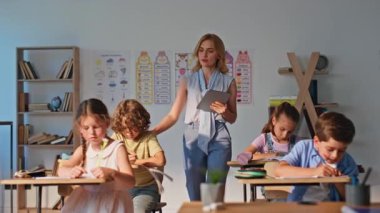 Grup çocukları ödev yaparken sınıf sıralarına oturuyorlar. Sınıfta öğrencilerin yazı yazmasını izleyen genç bayan öğretmen. İlkokul öğrencileri ders çalışıyor. Bayan eğitimci bilgi dikte ediyor
