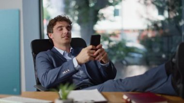 Rahatlatıcı iş adamı akıllı telefondan sosyal medyayı karıştırıyor özel kabinede bacakları masaya koyuyor. Mutlu adam cep telefonu ekranına bakıp ofise gülüyor. Gülümseyen CEO mesajlaşması iş molalarının keyfini çıkarın