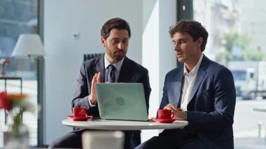 Profesyonel adamlar kapalı bir restoranda oturup projeyi tartışıyorlar. Gülümseyen iş arkadaşları modern kafeterya masasında dizüstü bilgisayarla iş birliği yapıyorlar. Kafedeki şirket toplantısında neşeli bir takım çalışması.