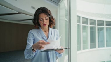 Belgeleri okuyan iş kadını ofis koridorunu kapatıyor. Odaklanmış profesyonel yönetici şirket koridorunda cam kenarında yürüyen evrak işlerini inceliyor. Kadın patron işi aceleye getirmeye çalışıyor.