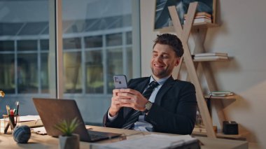 Modern ofis koltuğunda oturan neşeli profesyonel cep telefonu akşam kapanışı. Mutlu yönetici akıllı telefondan mesaj atıyor. Gülümseyen iş adamı cep telefonunda sosyal medyaya göz atıyor..