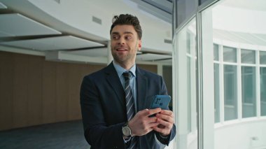 Gülümseyen yönetici ofis koridorundaki pencerelerde yürürken cep telefonu mesajı okuyor. Neşeli menajer molada akıllı telefonu kontrol ediyor. Rahat iş adamı koridorda cep telefonuna mesaj atıyor..