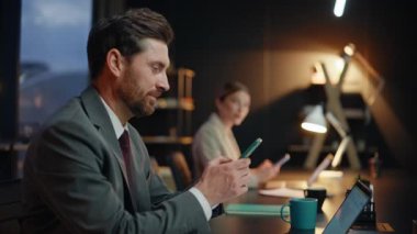 Akşam iş arkadaşları lamba ışığı çalışma alanındaki akıllı telefonları mesajlaşıyorlar. İki meslektaş birbirine gülümsüyor ve cep telefonlarıyla karanlık işyerinde oturuyor. Başarılı sakallı iş adamı cep telefonuna göz atıyor