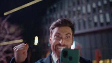 İş sahibi, akşam sokak portresindeki başarısına seviniyor. Heyecanlı duygusal direktör elinde cep telefonuyla harika haber mesajlarına tepki veriyor. Neşeli acemi adam el kol hareketi yaparak para kazanıyor.