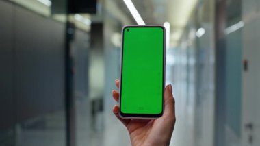 Boş koridorda el ele tutuşan, dikey model cep telefonu. Bilinmeyen bayan yönetici tek başına yeşil ekran akıllı telefon izliyor. İş kadını kapalı mekanda krom anahtar cep telefonu kullanıyor
