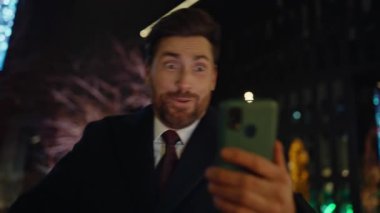 Karanlık mahalle portresinde komik bir patron çevrimiçi sohbet ediyor. Pozitif sakallı iş adamı akşam vakti şehrin yakın çekimlerinde sanal çağrıda eğleniyor. Duygusal adam sokakta el kol hareketi yapıyor. 