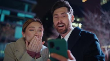 Çevrimiçi çift, gece şehir portresindeki video konuşmasında aşk jestleri gösteriyor. Mutlu kadın eşler hava öpücüğü gönderip akşam kentinde sanal sohbetin tadını çıkarıyor. Coşkulu çift çocuğu arıyor.