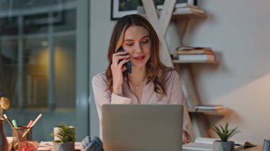 Akşam ofislerinde profesyonel iletişim cep telefonu danışmanlığı. Gülümseyen iş kadını kağıtları kontrol ediyor, masaları düzenliyor. Genç yönetici ticari akıllı telefon görüşmesi projesini tartışıyor