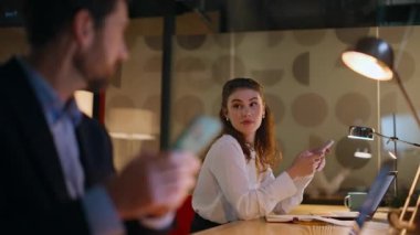 İki çalışan cep telefonlarını arıyor karanlık ofiste birlikte çalışıyorlar. Gülümseyen meslektaşlar akşam ofisindeki akıllı telefonlara göz atıyorlar. Mesaiye ara verip telefonları kullanan rahat iş arkadaşları..
