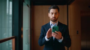 Zengin iş adamı cep telefonuna mesaj atıyor modern ofis koridorunda yürüyor. Kendine güvenen sakallı iş sahibi akıllı telefona mesaj gönderiyor. Lüks şirket binasında e-posta okuyan zarif girişimci..