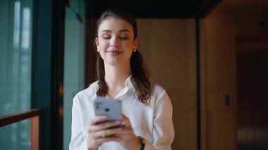 Akıllı telefondan yürüyen şirket binasının koridoruna mesaj atan memnun iş kadını. İş yerinde SMS okuyan mutlu bir iş kadını. Gülümseyen kız ofis koridorunda bankacılık uygulaması telefonuna bakıyor.