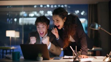 İki yönetici, akşam kurumsal iş yerinde dizüstü bilgisayar ekranında başarıya seviniyor. Heyecanlı meslektaşlarım duygusal tepki göstererek harika haberler okuyorlar. Coşkulu iş arkadaşları iş başarısının tadını çıkarıyorlar.