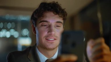 Akşam işadamı cep telefonuyla sosyal medyayı izliyor karanlık ofiste oturuyor. Geç saatte akıllı telefon mesajı okuyan gülümseyen adam. Mutlu genç işadamı cep telefonunda çevrimiçi içeriği tarıyor