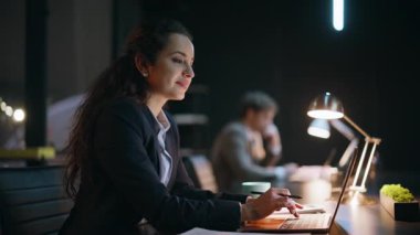 Gece ofiste çalışan gülümseyen iş kadını dizüstü bilgisayarı klavyeyle kapatıyor. Pozitif bayan yönetici, meslektaşı konuşuyor. Akşamları birlikte çalışmaktan mutlu. İçerideki bilgisayarlara mesaj atan neşeli kadın işçi.