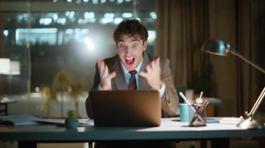 Heyecanlı iş adamı duygusal açıdan başarılı bir projeye tepki veriyor. Aşırı mutlu adam karanlık iş yerinde dizüstü bilgisayara bakıyor. Neşe dolu müdür, akşam odasında el kol hareketi yapıyor.