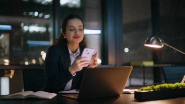 Karanlık iş yerinde cep telefonu mesajlarını okuyan gülümseyen bir iş kadını. Rahat bayan akıllı telefona göz atıyor ve gece ofisinde güzel mesajların tadını çıkarıyor. İçerde çalışan bir kadın yönetici e- postaya cevap veriyor