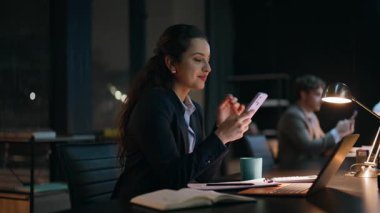 Geç saatte iş arkadaşları karanlık ofis kapanışında akıllı telefonlar kullanıyor. Cep telefonuna bakan mutlu iş kadını dolap masasında neşeli hissediyor. Gülümseyen esmer kadın, işyerinde çalışmaktan rahatlıyor. 