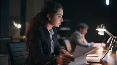 Odaklanmış yönetici, gece ofisinde evrak işleriyle uğraşıyor. Fazla mesai yapan bilgisayar ekran görüntüleme çalışanı karanlık çalışma mali raporlarını kontrol ediyor. Gülümseyen kadın kazanan lamba ışığı altında başarıyla seviniyor.