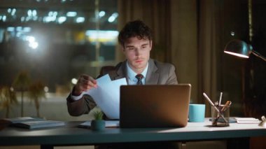 Odaklanmış yönetici, gece ofisinde evrak işleriyle uğraşıyor. Fazla mesai yapan bilgisayar uzmanı karanlık dairedeki mali raporları kontrol ediyor. Lamba ışığı altında sevinen belgeleri okuyan gülümseyen adam.