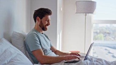 Uyanık iş adamı sabahın erken saatlerinde laptopta çalışıyor. Rahat adam rahat yatak odasındaki bilgisayarı kullanarak beyaz yatak odasına uzanıyor. Tembel evde geçen hafta sonu gülümseyen sakin esmer, dijital kapanış aygıtı