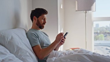 Sinirli adam sabahları hareketli görünüyor. Stresli uykulu erkek akıllı telefon kullanıyor. Gün ışığında cevapsız aramaları kontrol ediyor. Sinirli uyanık adam yatakta uzanıp sosyal medyayı okuyor ve mesajları tek başına yazıyor. 