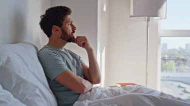 Hüzünlü adam, modern otel odasında yatarken mutsuz oluyor. Üzgün adam rahat yatak odasında rahat rahat düşünceli bir şekilde dinleniyor. Erkek esmer, sabah güneşinde beyaz battaniye içinde dalgın bir ifade takınıyor. 