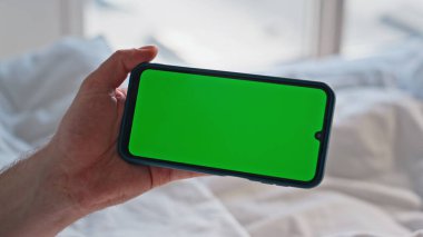 Hafta sonu yakın çekimde online alışverişleri seçen bir adam el kamerasıyla cep telefonunu yatakta kaydırıyor. Kimliği belirsiz adam rahat ev yatağında yeşil ekran akıllı telefon kullanıyor. Krom anahtarlı erkek cep telefonu 