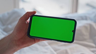 Açık ev yatak odasındaki yeşil ekranlı cep telefonuna dokunuyor. Kimliği belirsiz bir adam, beyaz bir yatakta krom anahtarlı akıllı telefon tutuyor. Dairedeki adamın el taraması modeli cep telefonu uygulaması 