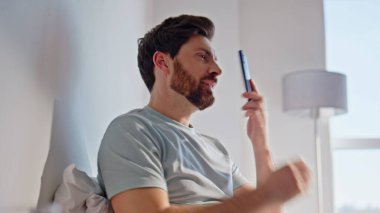 Rahat, esmer, konuşan cep telefonu, rahat yatak lambası dairesinin yakınında. Neşeli adam rahat yatak odasında akıllı telefonu arıyor. Gülümseyen adam hafta sonu evde iletişimin keyfini çıkarıyor. 