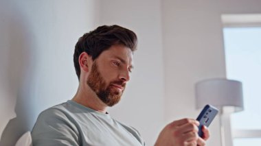 Mutlu esmer cep telefonları. Yatakta rahat bir şekilde uzanıyorlar. Akıllı telefon uygulamasına göz atan neşeli bir adam hafta sonu sabah yatak odasında online sipariş veriyor. Rahatlamış adam eve gülümseyen cep telefonu ekranı 
