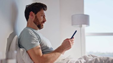 Hafta sonları cep telefonuna göz gezdiriyorum. Tasasız adam beyaz yatak odasında oturan akıllı telefon ekranına bakıyor. Rahatlamış esmer, yatak odasında cep telefonu tutuyor ve sükunetin tadını çıkarıyor.