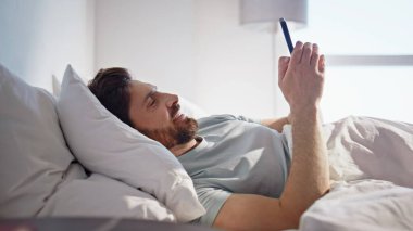 Mutlu adam yatakta akıllı telefon ekranı gibi görünüyor. Uykulu erkek hücrede yatan mesajı okur. Yakışıklı sakin adam sabah cep telefonu kullanarak kontrol et. Uyanmış neşeli esmer rüya İyi haberlerin tadını çıkar