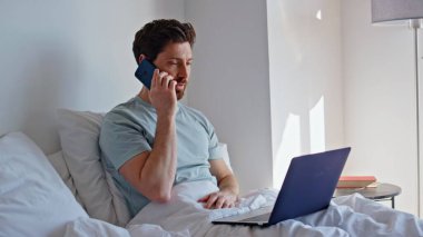 Otel odasında dizüstü bilgisayarla cep telefonu oturmaktan bahseden olumlu bir yönetici. Neşeli, duygusal bir adam cep telefonuyla iş konuşuyor. Gülümseyen bir çalışan yatak odasından çalışıyor.