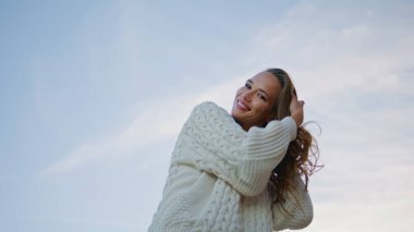 Gülümseyen bir model, saçlarını ayarlıyor. Akşam ufuk çizgisine bakıyor. Açık gökyüzünün önünde beyaz süveter giyen mutlu, esmer bir kamera. Kaygısız kadın doğa alacakaranlığının keyfini çıkarıyor.