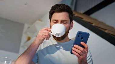 Sabahçı adam cep telefonundan mesaj okuyor. Ev mutfağında kahve içiyor. Yakın plan. Mutlu adam akıllı telefonu kaydırıyor ve sessiz kahvaltının tadını çıkarıyor. Modern mutfakta rahatlamış erkek aromatik içeceği yudumluyor.