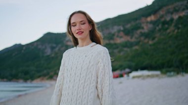 Kumralın tadını çıkaran mutlu esmer, yakın plan çekimde. Kumlu deniz kıyısında rahat bir süveter giyerek poz veren çekici bir model. Sahilde duran gülümseyen kadın yeşil dağları çevreledi..