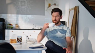 Sabah mutfağında çay içen dalgın bir adam. Rahat adam sıcak içeceğini yudumluyor ve sakin atmosferin tadını çıkarıyor. Düşünceli esmer adam pencereye bakıyor. Hafif dairede kahve molası veriyor.
