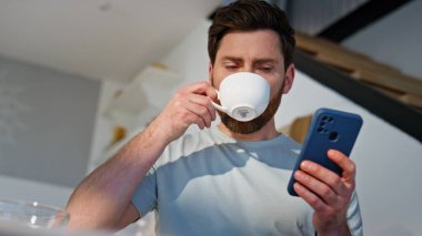 Sabahçı adam akıllı telefonları karıştırıyor. Mutfağı kapatıyorlar. Yakışıklı, aromatik kahve içen cep telefonu görünümlü bir adam. Dairede cep telefonu uygulaması kullanarak rahatlamış erkek alışverişi