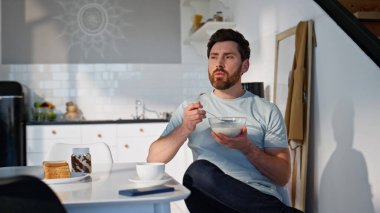 Aç esmer kahvaltı yiyor modern mutfakta yakın plan oturuyor. Tıraşsız, gülümseyen ve gevreğin tadını çıkaran adam ışıklı evin camına bakıyor. Sakin serbest çalışan öğle yemeğini evde tek başına yiyor. 