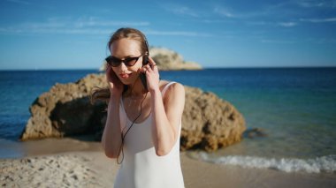 Plajda dans eden kulaklıklı kadın güneş gözlüğü takıyor. Gülümseyen hippi kız güzel okyanus dalgaları karşısında müzik dinliyor. Mutlu bayan güneşli kumlu sahilde dinleniyor. İyi tatiller.