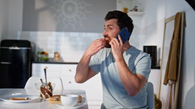 Kahvaltılık adam, akıllı telefondan arayıp mutfakta sandviç yiyor. Evde oturan rahat adam cep telefonuyla sabah keyfine bakıyor. Sakin erkek rahat evde telefonda haberleri tartışıyor.