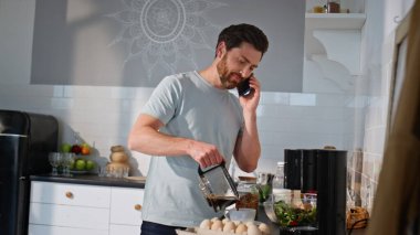 Çağrı adam kahve doldurarak konuşuyor kapalı mutfakta akıllı telefon konuşarak. Apartman mutfağında cep telefonuyla sabah içeceği hazırlayan pozitif bir adam. Rahatlamış erkek kahvaltıda telefon haberlerini tartışıyor. 