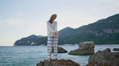 Rüzgarlı plajda poz veren turist üşüyor. Okyanus kıyısında dalgalı saçlarıyla çıplak ayakla kayaların üzerinde duran mutlu kadın manken. Huzurlu bayan tek başına alacakaranlık deniz manzarasının tadını çıkarıyor. Deniz seyahati konsepti.