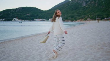 Kaygısız kız kumsala adım atıyor ve haftasonunu sahilde geçiriyor. Gülümseyen kadın turist güzel okyanus kıyısında çantasıyla yürüyor. Mutlu tatilde sabah deniz manzarasının tadını çıkaran neşeli bir kadın.