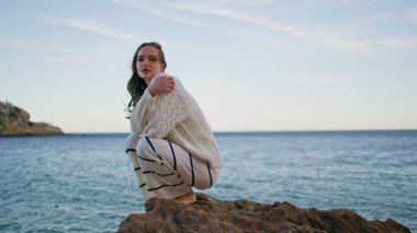 Sahildeki kayalıklarda çömelmiş deniz manzaralı güzel bir model. Sıcak kazaklı romantik kadın okyanus kıyısında rüzgarlı bir akşamın tadını çıkarıyor. Huzurlu hayalperest bayan tatilde dalgalı deniz yüzeyinde dinleniyor..